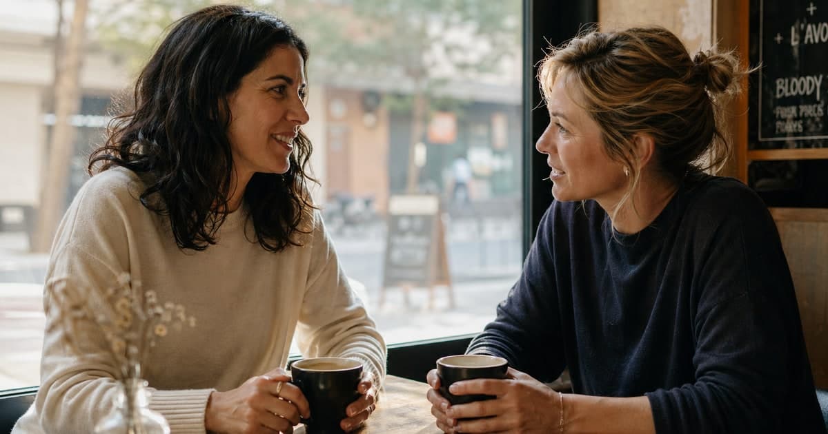 Two friends in their late 30s sitting across from each other at a sunlit cafe table, mid-conversation, hands cradling coffee cups. Editorial photograph, warm natural light, candid framing.