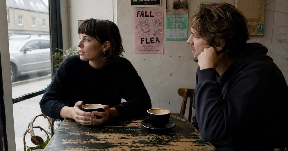 Candid photo: two people at a small cafe table mid-conversation, one glancing toward the window, worn tabletop with two coffee cups, local flyers on the wall behind them, overcast natural light.
